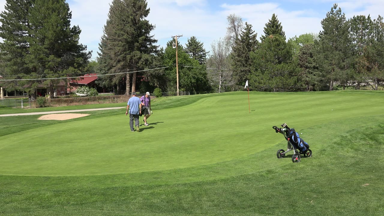 Two adult men are closer together on the Denver course, with one near a golf bag and push cart while the fairway, bunker, trees, and flag remain visible.