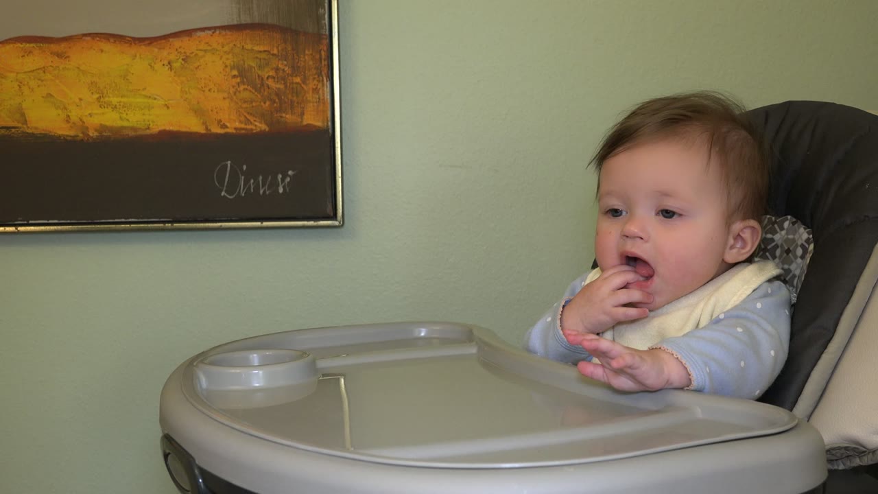 The baby remains seated in the high chair with mouth open, one hand near the face, and the tray and wall art visible in the Westminster home interior.