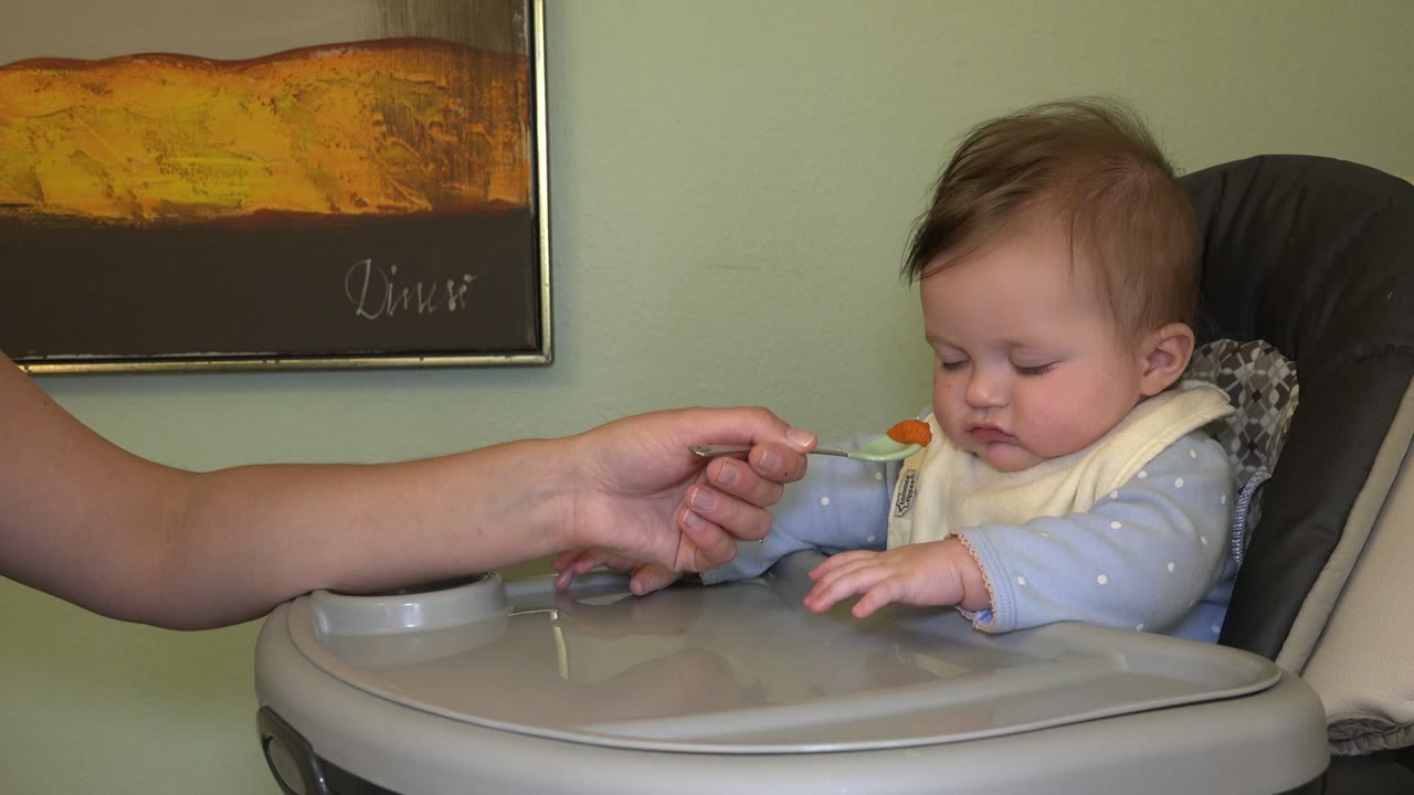 Baby in a high chair looks down as an adult offers a spoon.