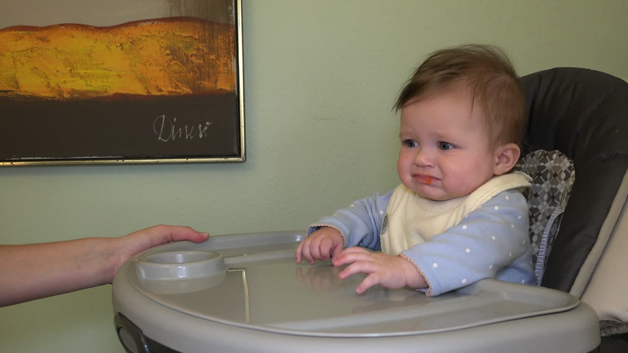 At 0:00 in Westminster, Colorado, a baby sits in a high chair with food on the face as an adult hand reaches near the gray tray from the left.