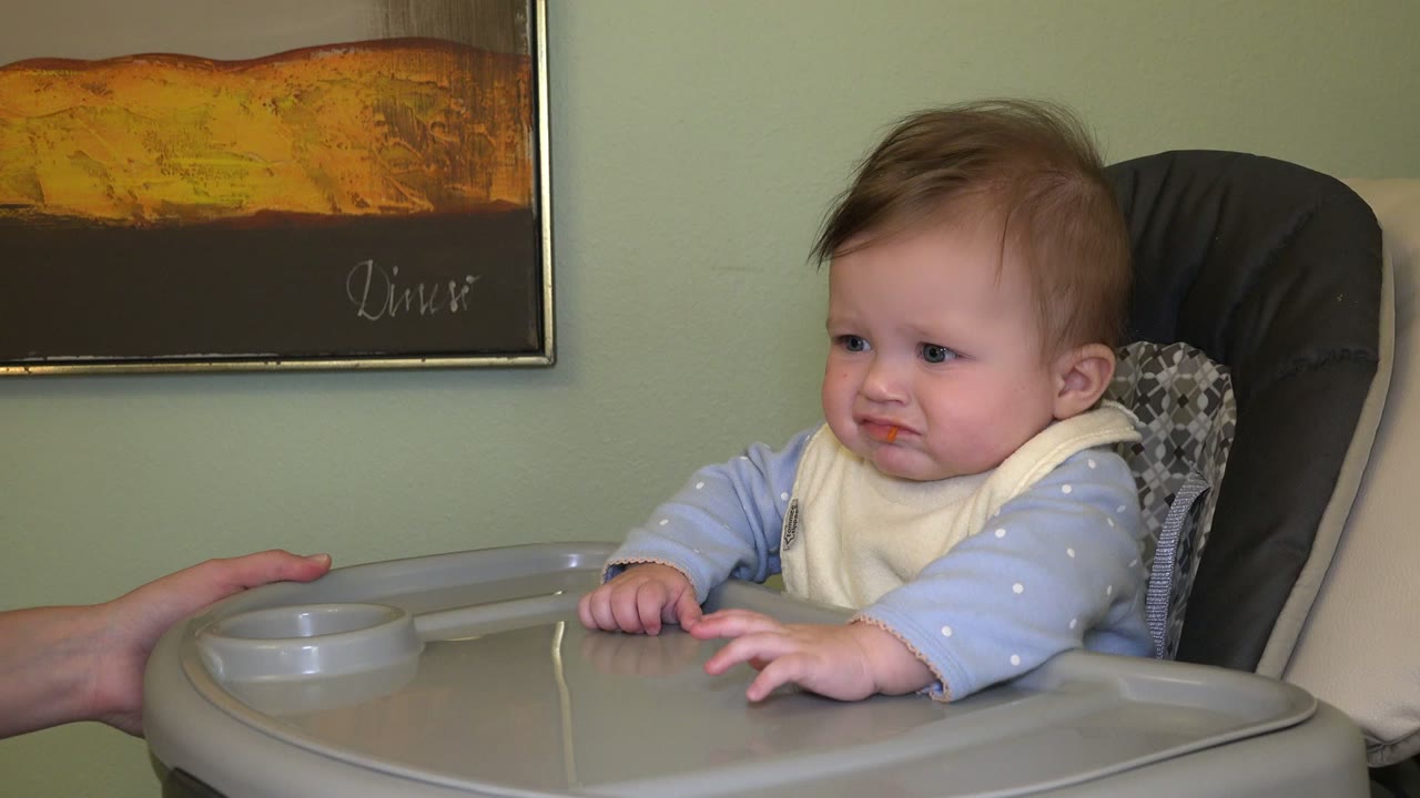 At 0:01 in Westminster, Colorado, the baby leans forward with both hands near the tray while the adult hand remains at the left edge of the frame.