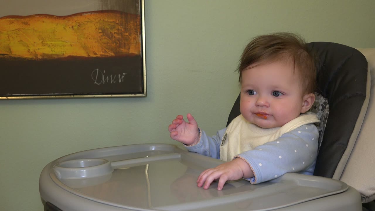 At 0:01 in Westminster, Colorado, the baby remains centered in the high chair, looking forward with the gray tray in front and framed wall art behind.