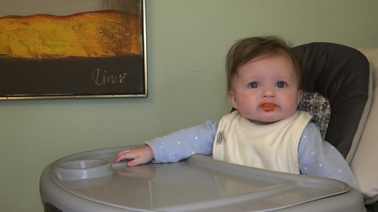 A baby sits in a gray high chair in Westminster, Colorado, facing the camera with orange food on the mouth and framed wall art behind.