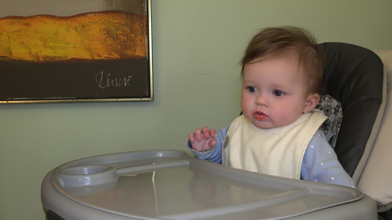 The baby remains in the Westminster, Colorado highchair setup, turning attention forward with one small hand lifted above the gray tray.