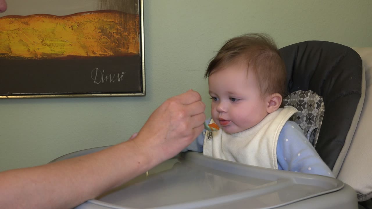 Adult hand brings a spoon toward a baby in a highchair.