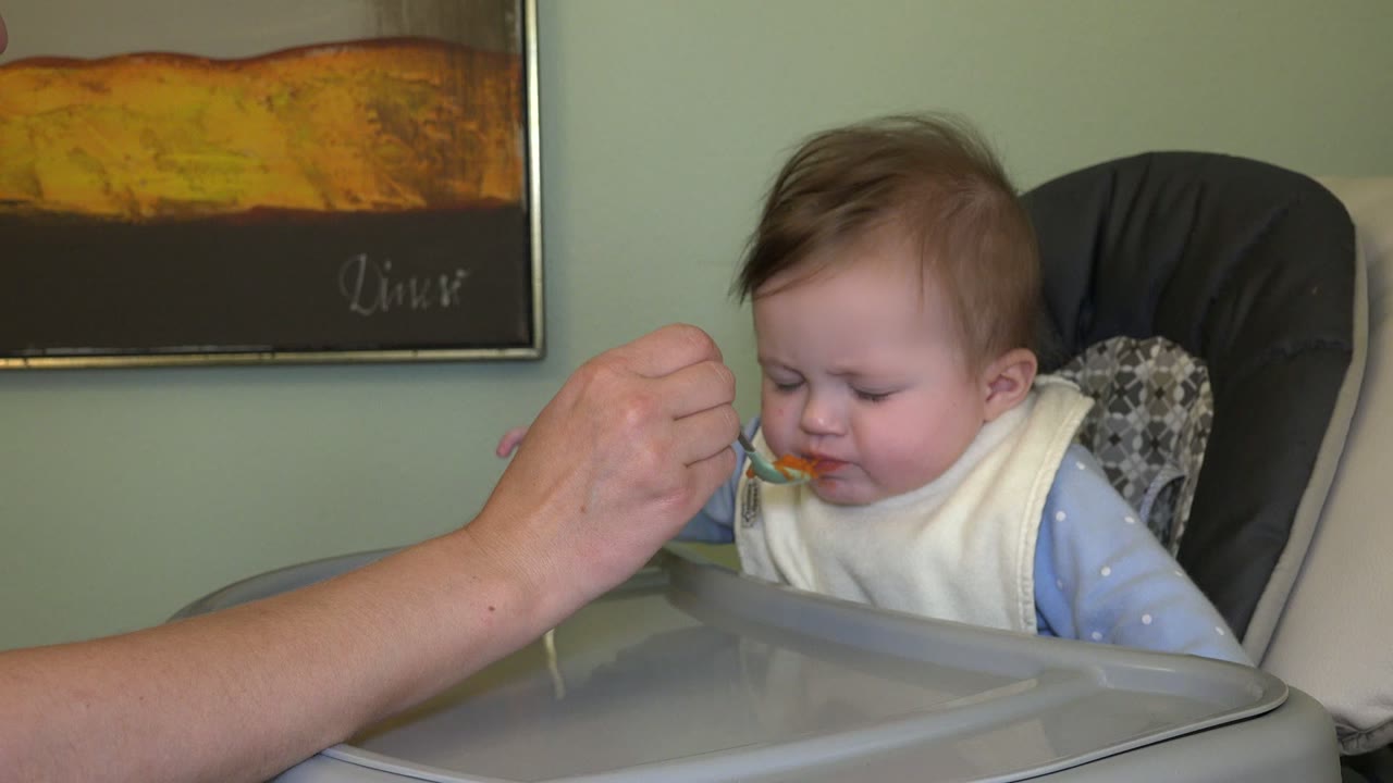 In Westminster, Colorado, the baby closes their eyes while an adult holds a spoon of orange food at the baby's mouth in the highchair.