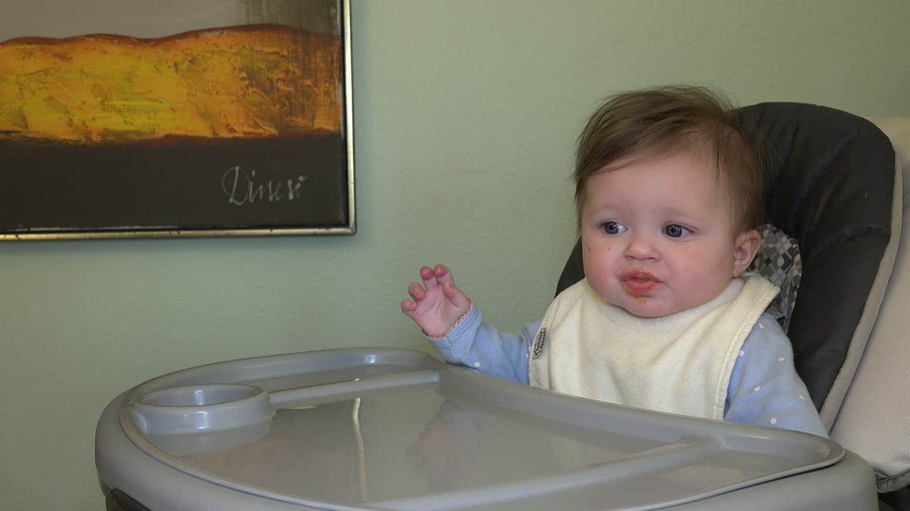 Baby in a high chair with a tray and wall art behind.