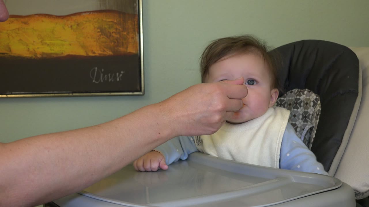 In Westminster, Colorado, an adult arm crosses the frame as a spoon is placed near the baby's mouth in the high chair.