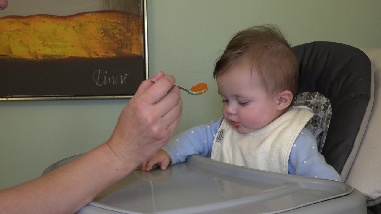 At 0:01 in Westminster, Colorado, the spoon of orange puree remains raised in the foreground while the baby sits back in the high chair wearing a white bib.