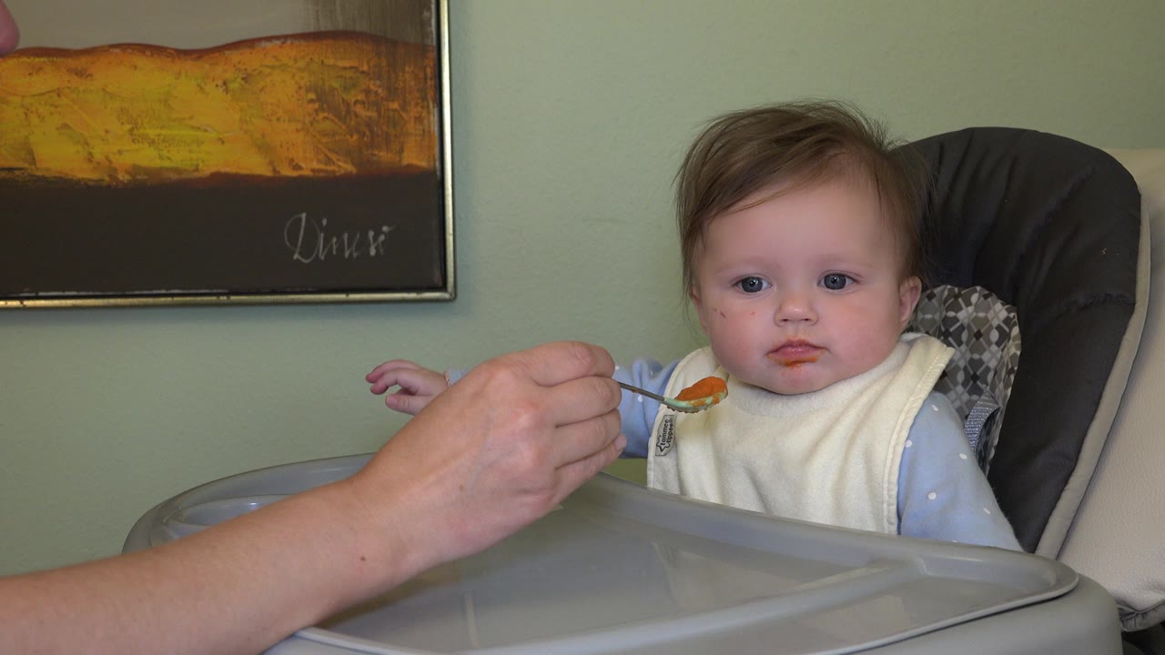In Westminster, Colorado, a baby sits in a high chair as an adult hand holds a spoonful of orange food near the baby's mouth.