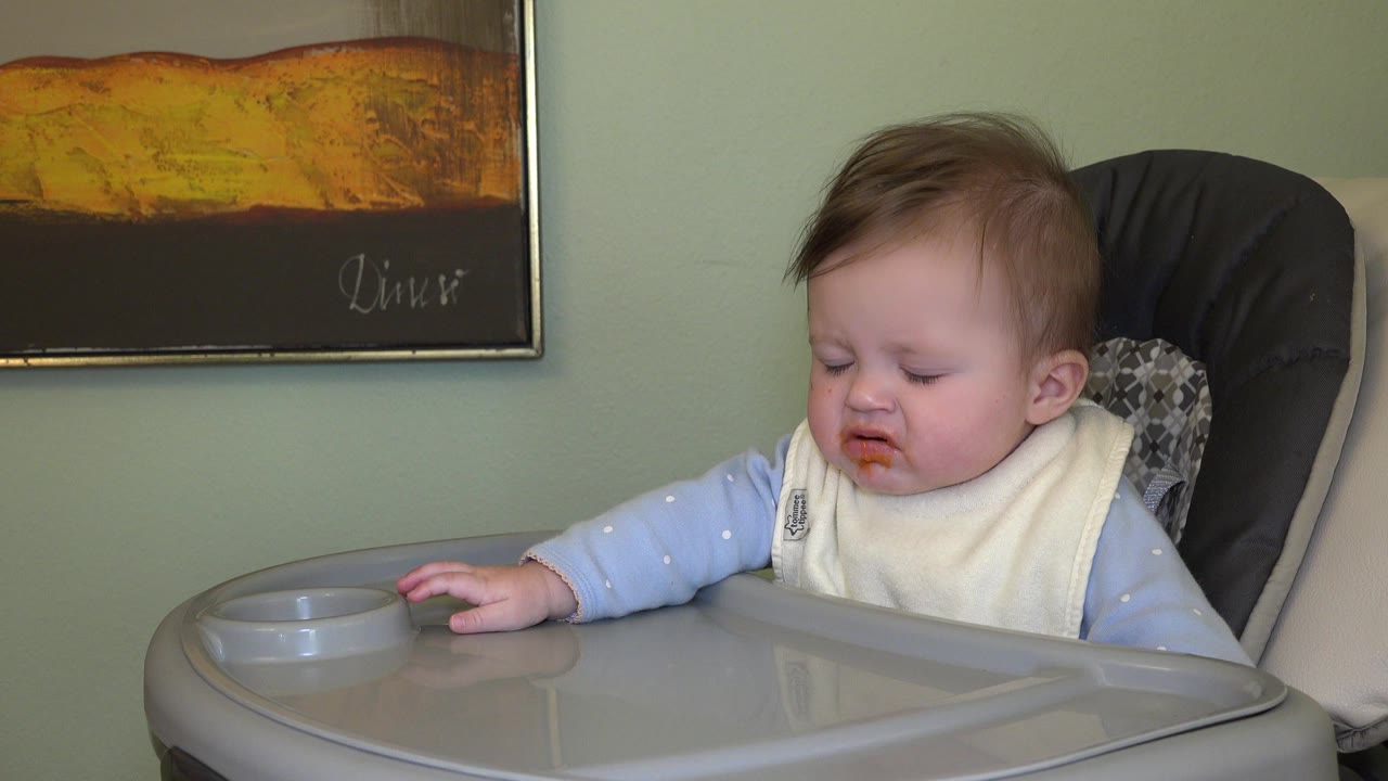 The Westminster, Colorado baby looks down with one hand near the tray edge, wearing a bib in the same indoor high-chair setup.