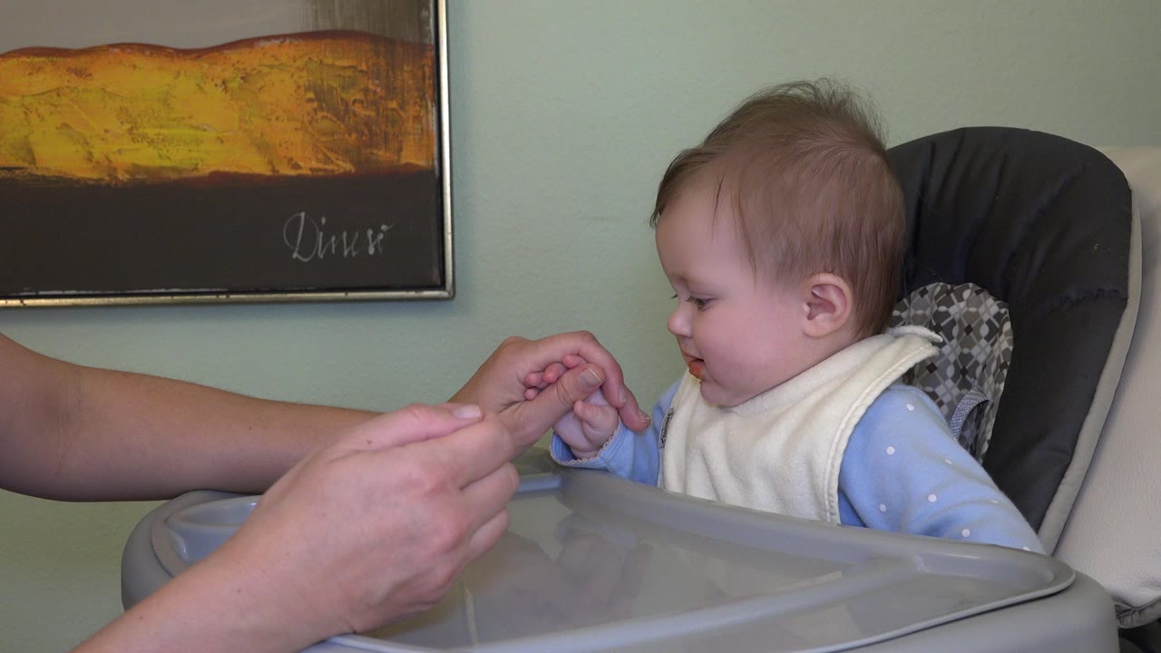 Baby in a high chair reaches toward an adult hand.