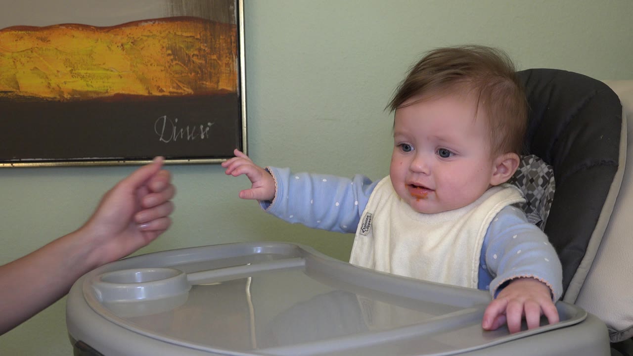Baby in a high chair reaches toward an adult hand.