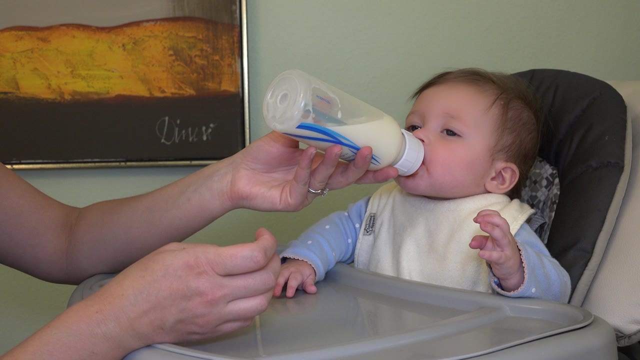 In Westminster, Colorado, a baby girl in a high chair drinks from a bottle while an adult hand supports it near a gray tray and framed wall art.