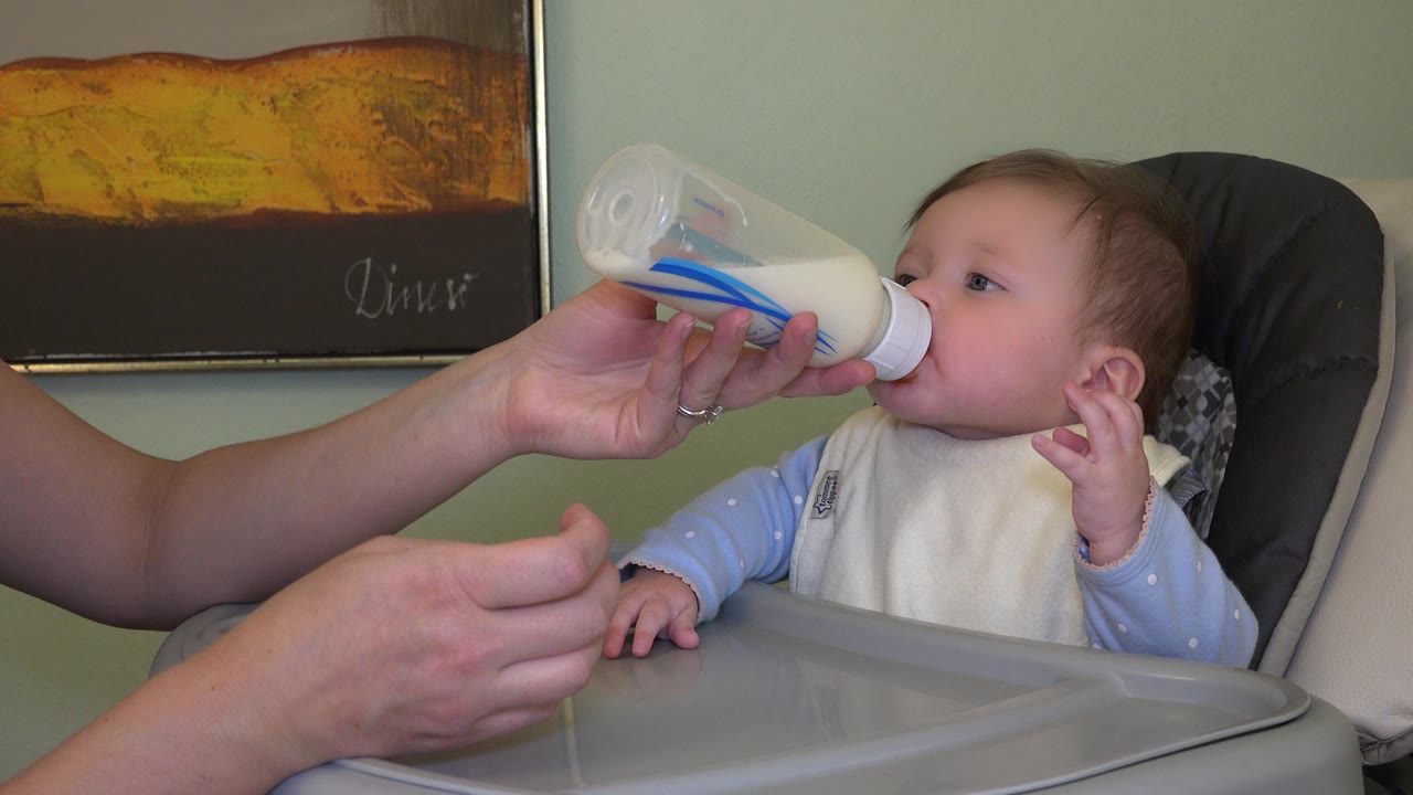 The baby drinks from the bottle and lifts a hand beside her face, with the adult hand holding the bottle steady in the Westminster, Colorado interior.