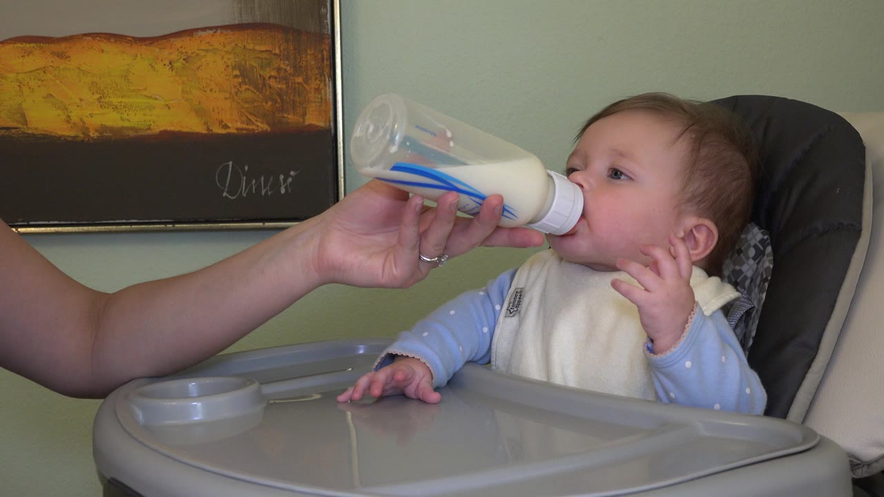 Baby in a high chair drinking from a bottle held by an adult hand.