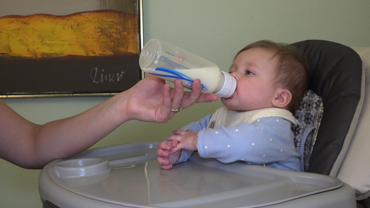 Baby in a high chair drinks from a bottle held by an adult hand.
