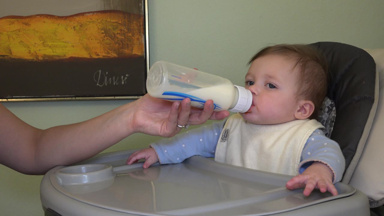 Baby in a high chair drinking from a bottle held by an adult hand.
