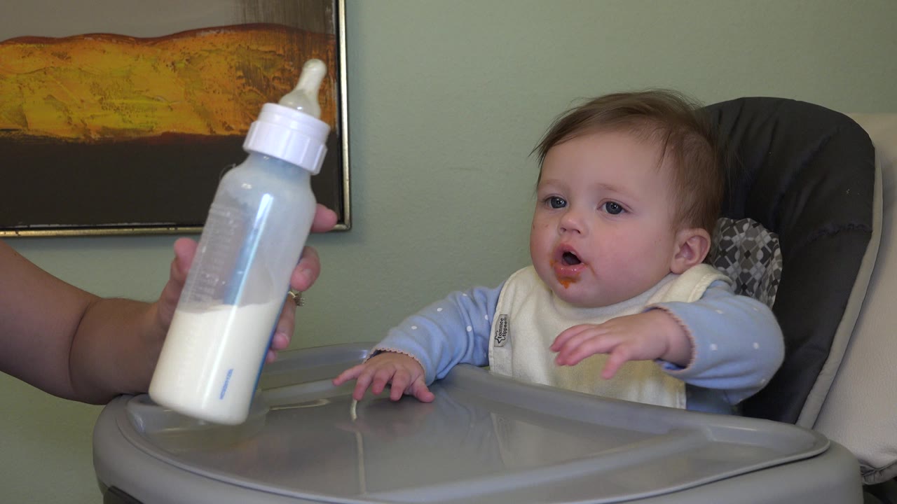 The baby remains seated in the high chair in Westminster, Colorado, with both hands raised while the bottle is held closer in the left foreground.