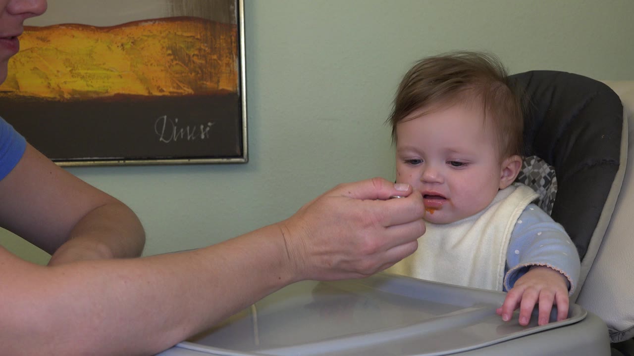 Father holds a spoon of orange puree near a baby in a high chair.