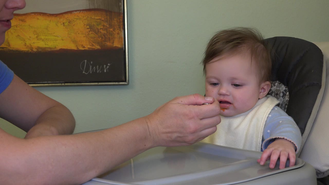 In Westminster, Colorado, the baby looks toward the spoon of orange puree while the father's arm reaches in from the left beside the high chair tray.