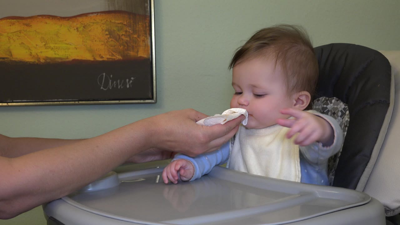 In Westminster, Colorado, the baby remains seated in the high chair and reaches one arm outward while the adult hand stays at the baby's mouth.