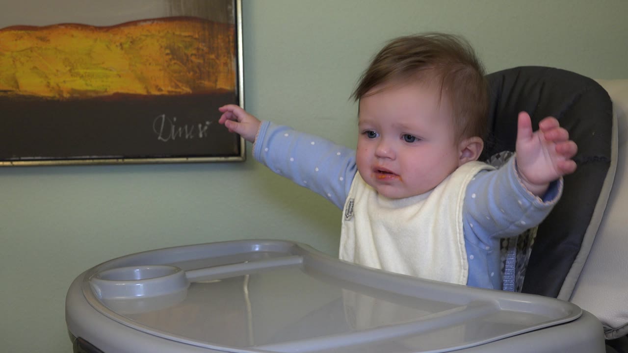 The baby in the Westminster, Colorado home setting lifts both arms above the high chair tray, with the framed wall art behind.