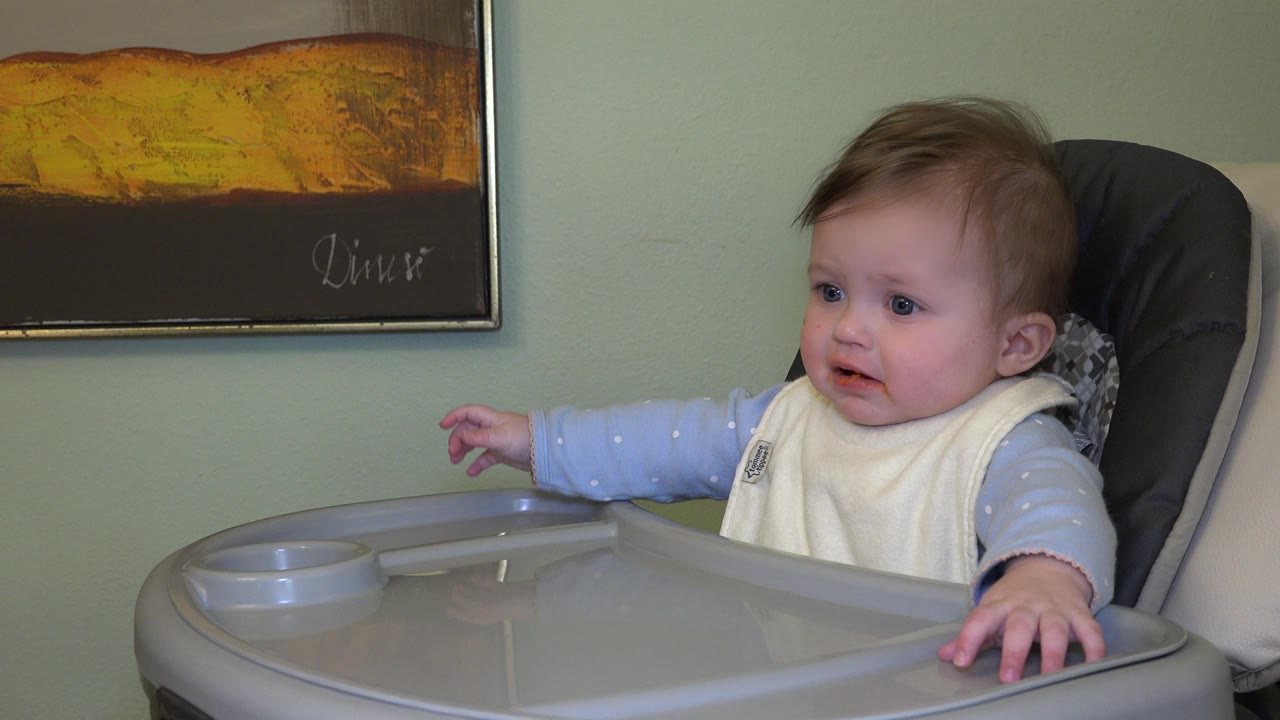 Baby in a high chair looking left with one arm extended.