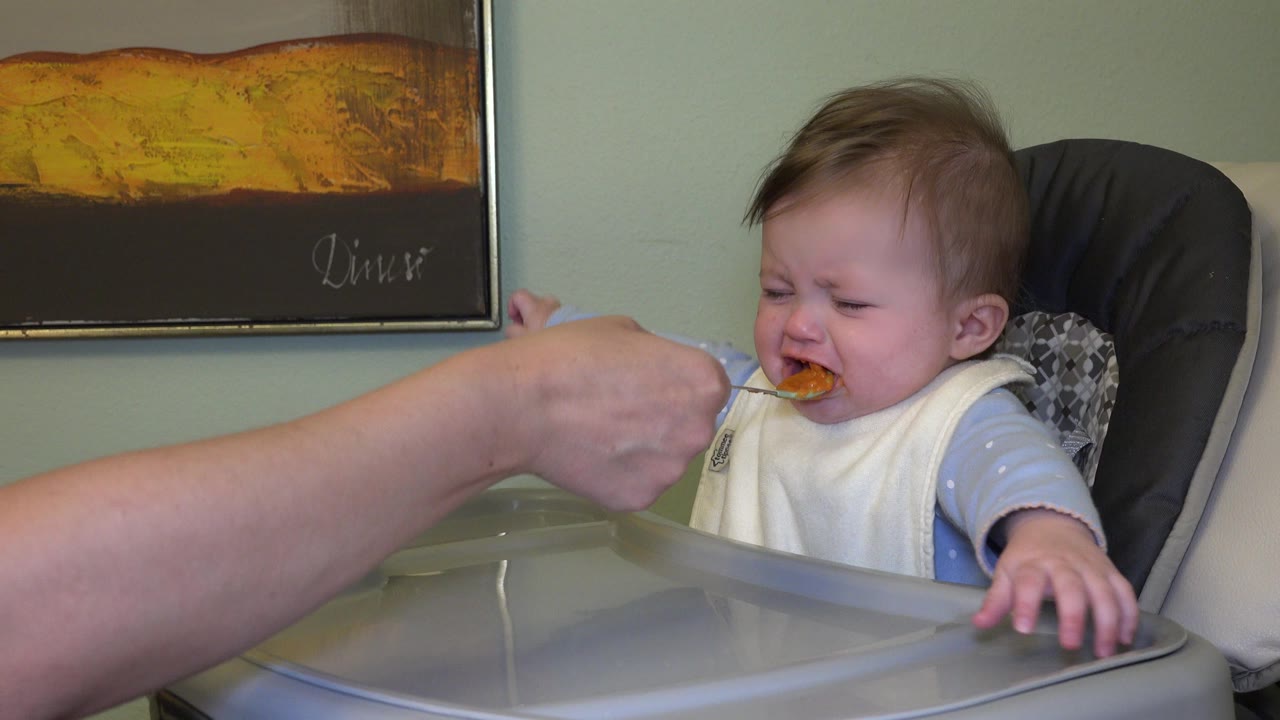 Adult hand offers mashed food to a crying baby in a high chair.
