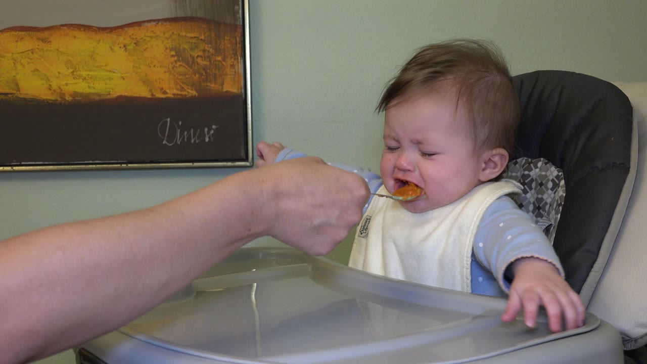 At 0:01 in Westminster, Colorado, the spoon reaches the baby's mouth while the baby squeezes their eyes and grips the high-chair tray.