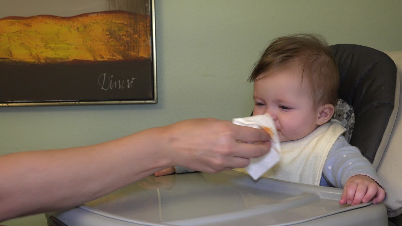 Adult hand wipes an infant's mouth in a high chair.