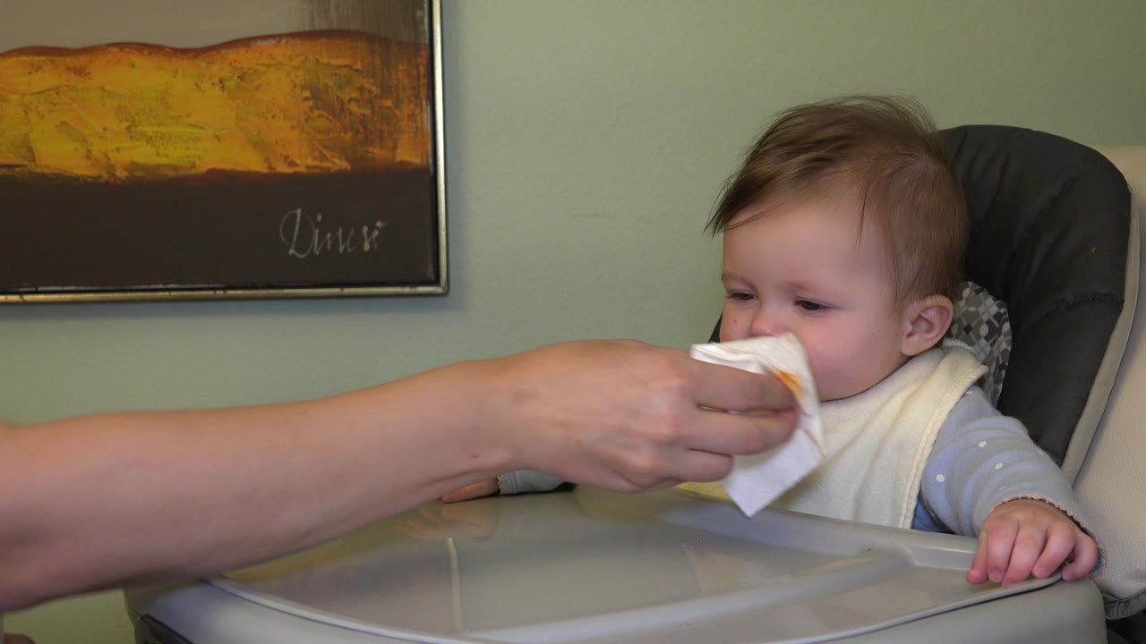 In Westminster, Colorado, the infant looks toward an adult's hand holding a white cloth at the child's mouth, with the high chair tray filling the foreground.