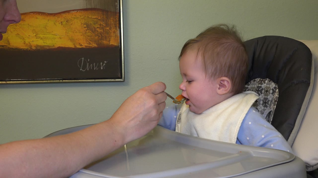 The toddler leans toward the spoon as the adult hand holds orange food close to the mouth, with the high chair tray filling the lower foreground.