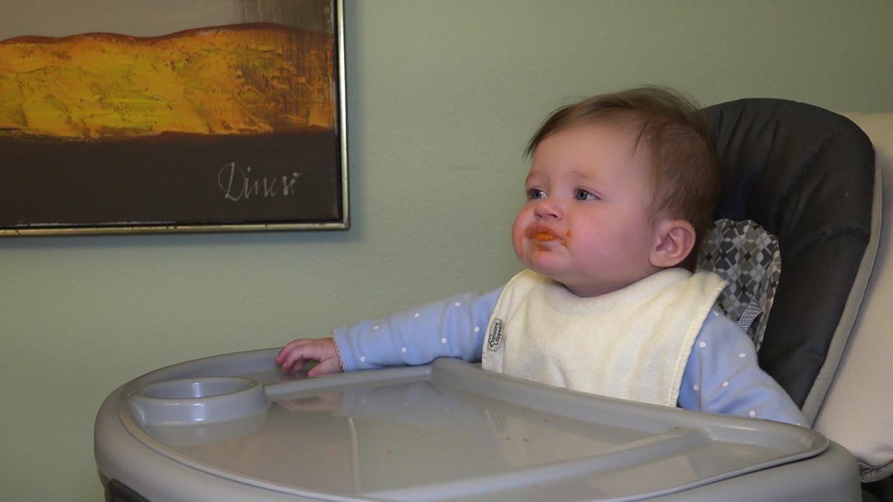 The baby turns attention toward a partial adult face entering from the right, with the gray tray and framed wall artwork still visible in the Westminster home interior.