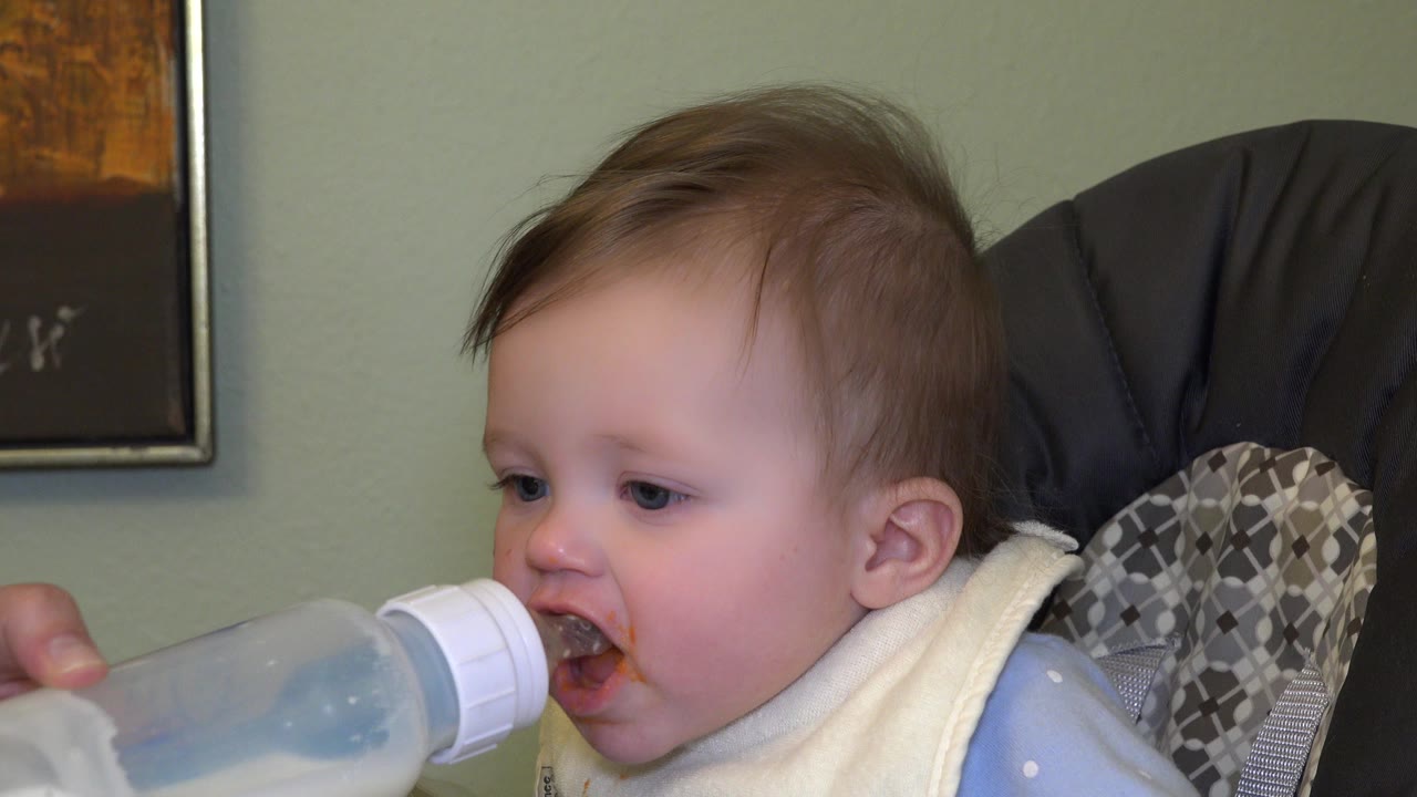 In Westminster, Colorado, a baby wearing a pale bib sits indoors as a bottle nipple is held near the mouth, with a green wall and framed artwork behind.
