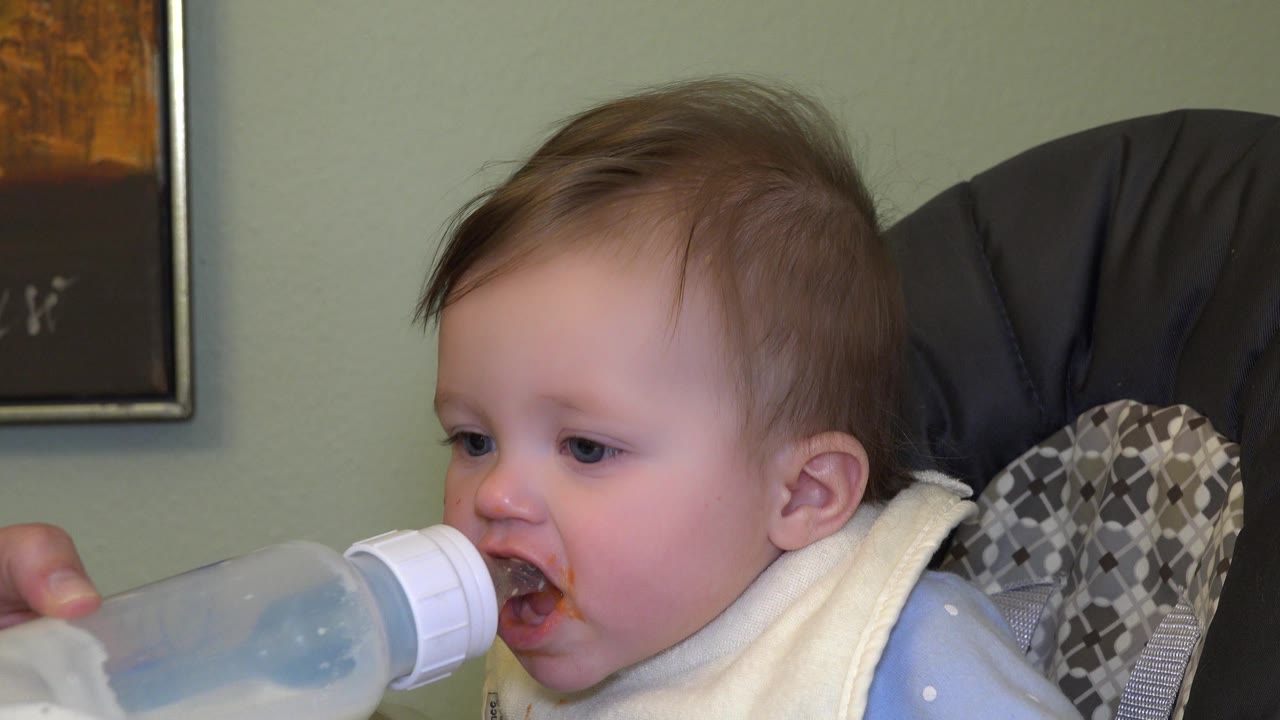 In Westminster, Colorado, the baby opens the mouth toward the bottle while an adult hand holds it from the left side of the close indoor frame.