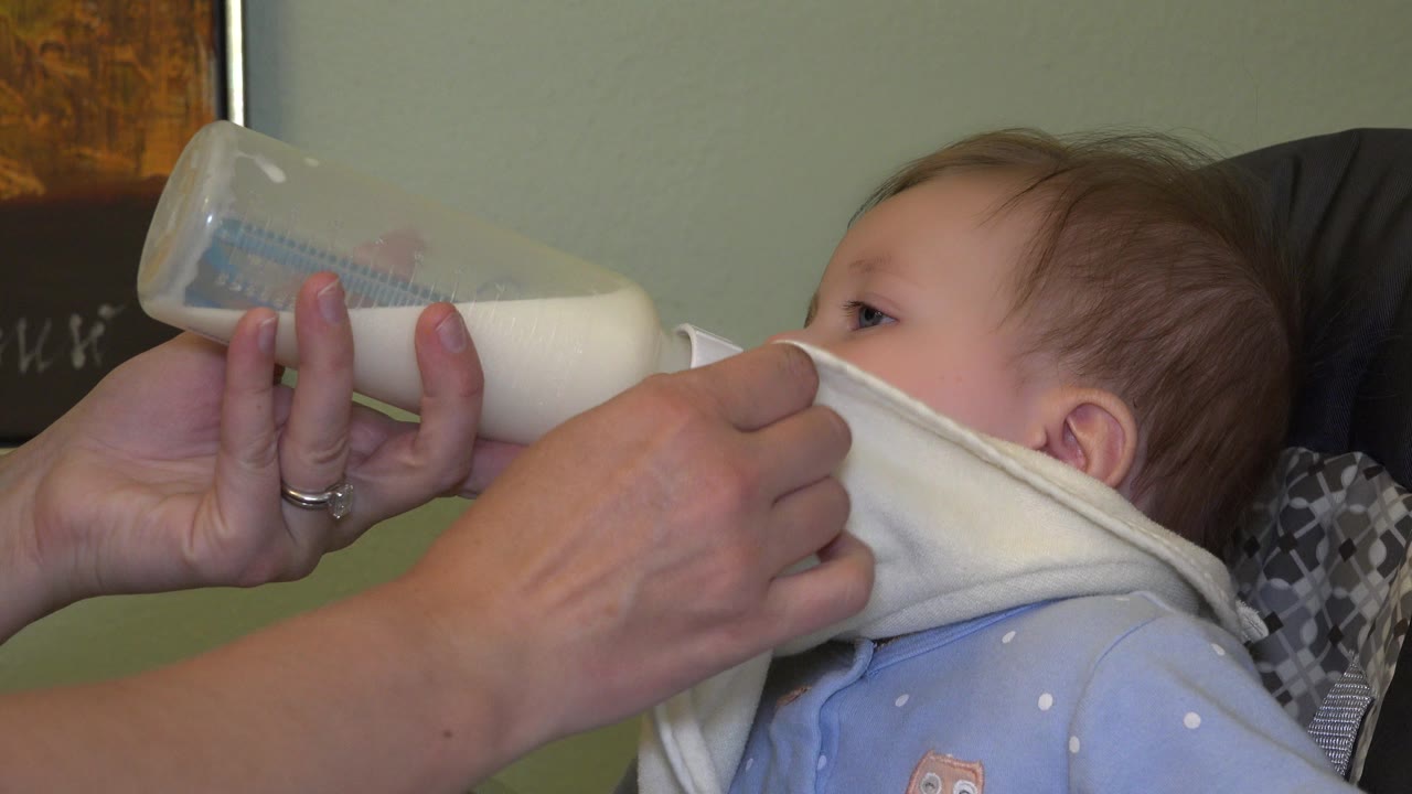 Adult hand holds a milk bottle to a baby wearing a bib.