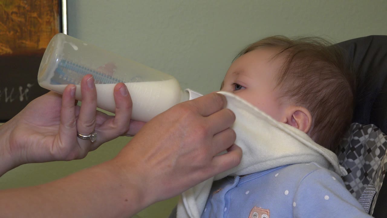 The baby continues drinking from the bottle as adult fingers support the nipple and bib, with the child's eye and cheek partly visible in the close frame.