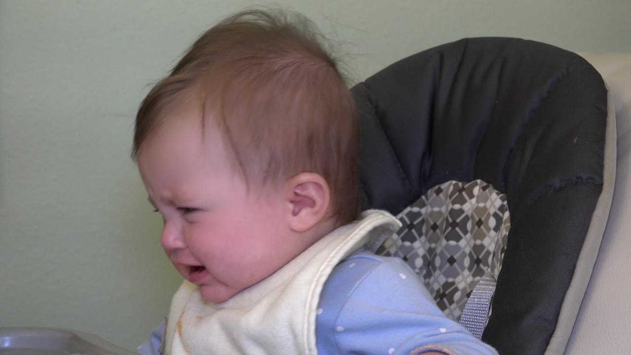 At 0:00, a reddish-haired baby in Westminster, Colorado cries with eyes squeezed and mouth open while seated in a padded child seat against a green wall.