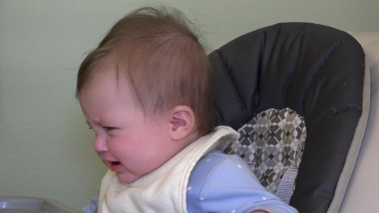 At 0:01, the baby remains turned slightly left, crying beside the patterned gray cushion and dark chair back in the Westminster, Colorado home footage.