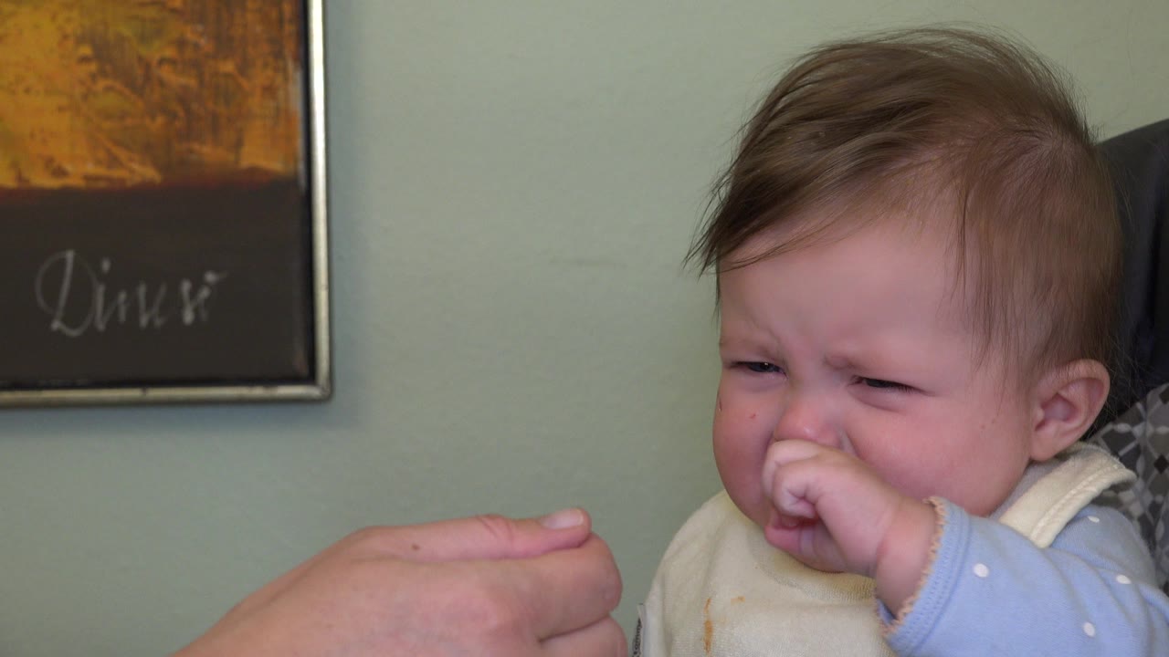 In Westminster, Colorado 2015 home footage, a baby in a bib cries with fingers near the mouth while an adult hand holds a spoon at the left edge.
