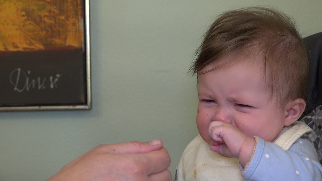 The baby remains teary with fingers pressed to the mouth as the caregiver's hand and spoon stay in the left foreground inside the Westminster, Colorado interior.