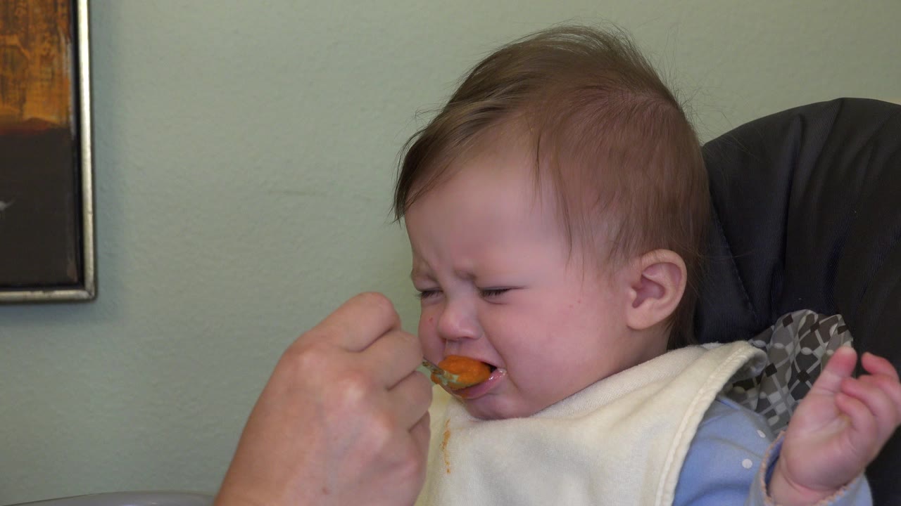 Baby grimaces as a spoon reaches their mouth.