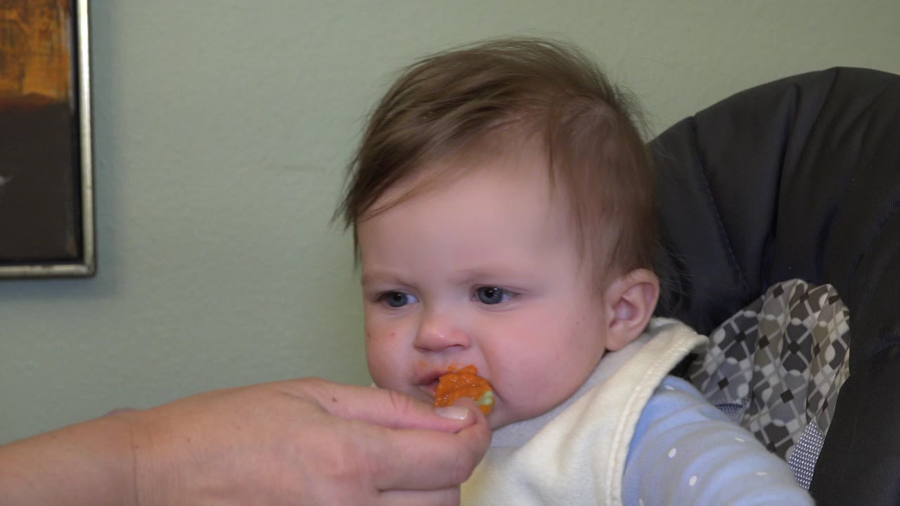 In Westminster, Colorado, the baby continues tasting orange mashed carrots while seated close to the camera in a patterned high chair.