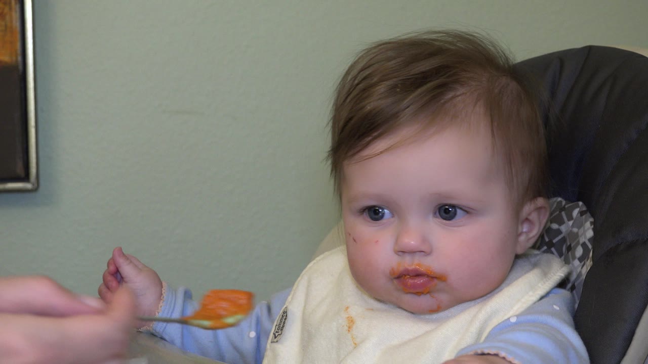 Child in a high chair faces an approaching spoon of orange food.