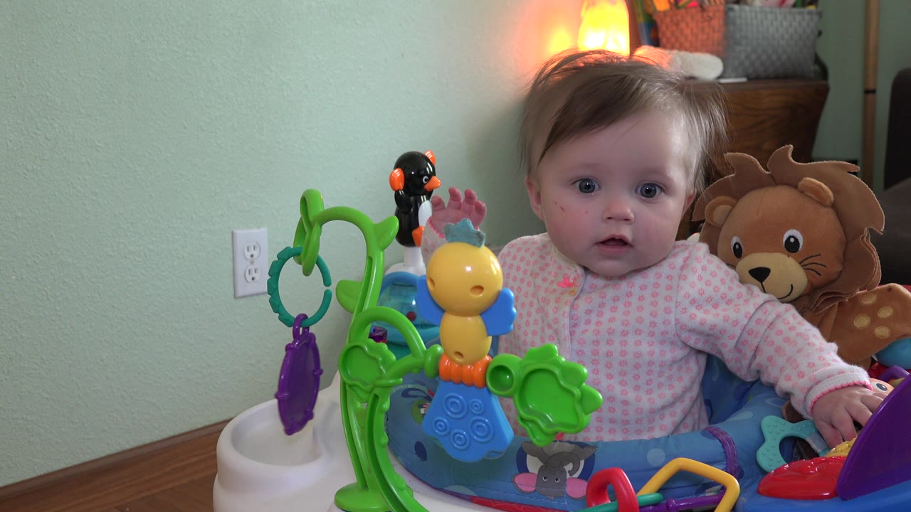 At 0:02, the baby girl sits slightly right of center in the walker, with the colorful toy cluster blocking part of her torso and the plush lion visible over her shoulder.