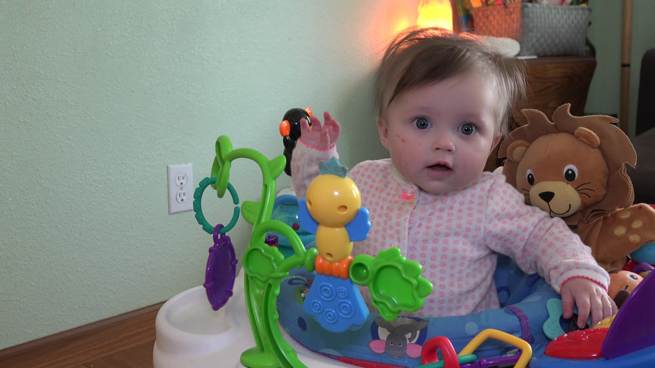 Baby girl raising one hand while seated in walker.