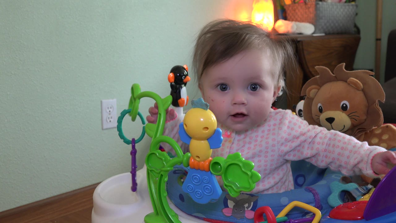 In Westminster, Colorado, the baby remains centered in the walker while a yellow toy and green play structure fill the foreground and plush animals sit near a dresser behind.