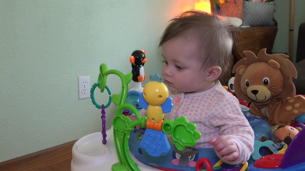 Baby seated in a colorful activity chair beside hanging toys.