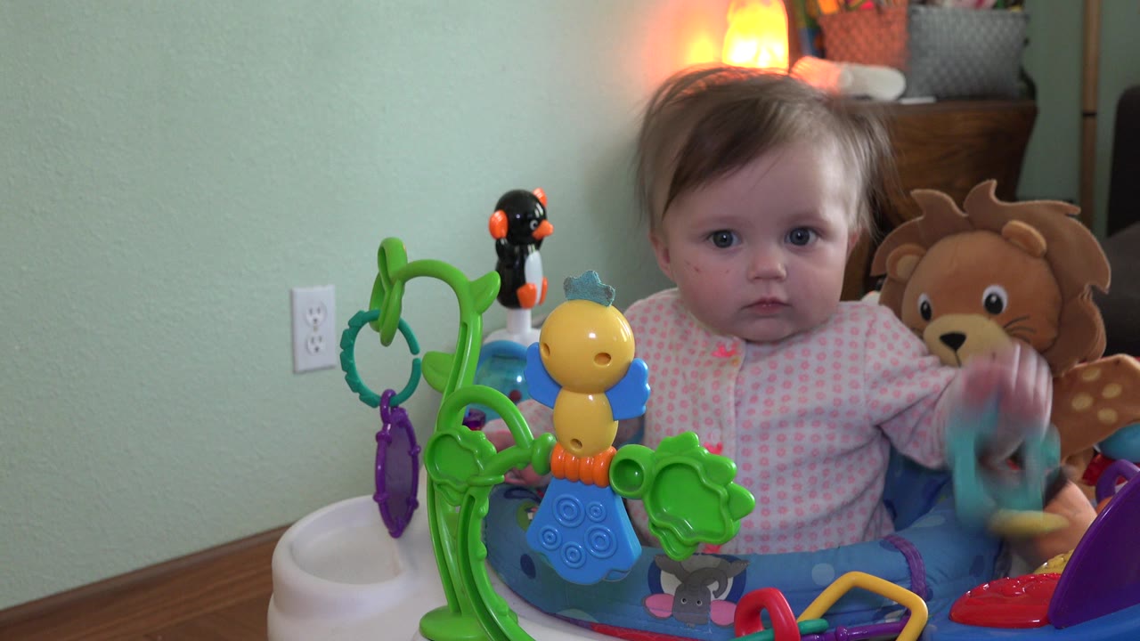 At 0:01 in Westminster, Colorado, the baby remains centered behind the activity toys while one hand lifts a small object near a red and purple toy area.